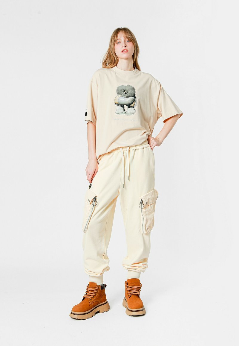 Camiseta oversized beige con estampado gráfico, combinada con pantalones cargo de crema clara que incluyen cremalleras. Botas marrones con cordones y suelas gruesas.