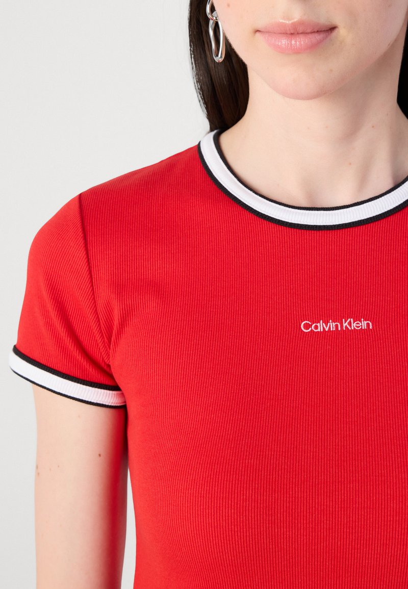 Camiseta de ribete rojo con mangas cortas, que presenta un cuello a rayas blancas y negras y un logotipo blanco de "Calvin Klein" en el pecho.