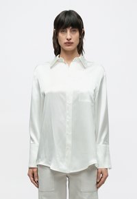 Bluza albă din satin cu guler clasic, nasturi în față și buzunar la piept. Mâneci lungi cu manșete late și tiv curbat.