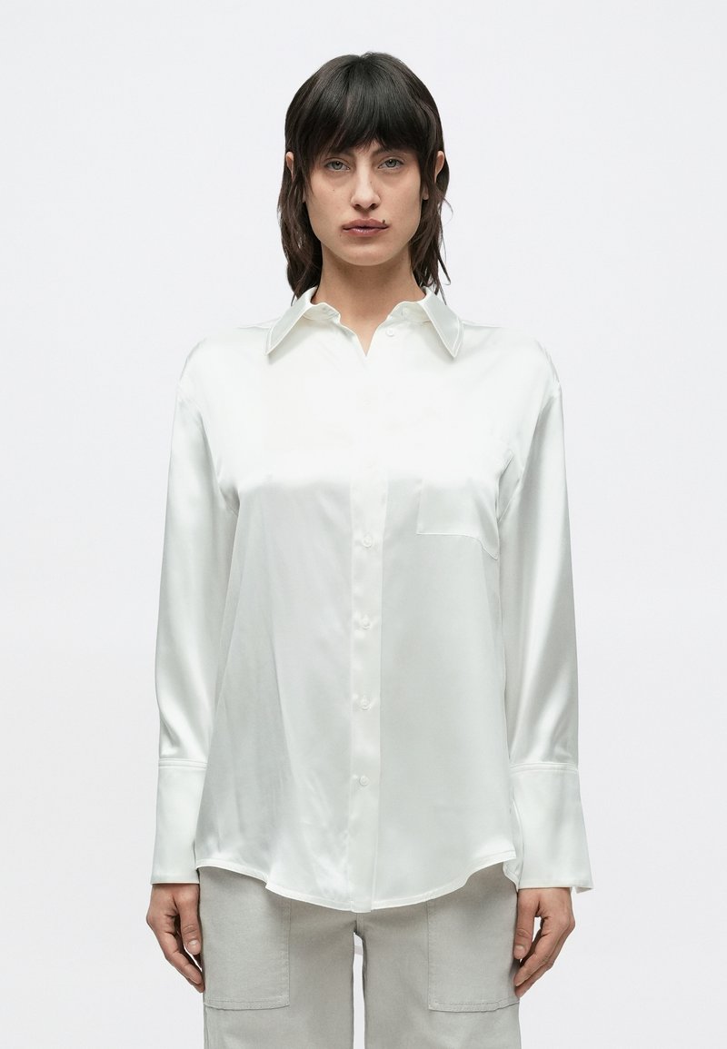 Bluza albă din satin cu guler clasic, nasturi în față și buzunar la piept. Mâneci lungi cu manșete late și tiv curbat.