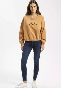 Musztardowa bluza z ribbowanym dołem i mankietami, z nadrukowanym tekstem. Zestawiona z ciemnymi dżinsami i białymi sneakersami. Gładka tekstura materiału.