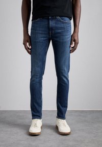 Man med slim-fit mörkblå jeans och vita sneakers som står på ett grått golv mot en ljusgrå vägg.