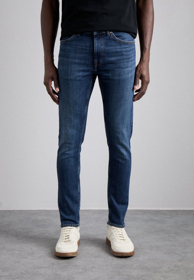 Man med slim-fit mörkblå jeans och vita sneakers som står på ett grått golv mot en ljusgrå vägg.