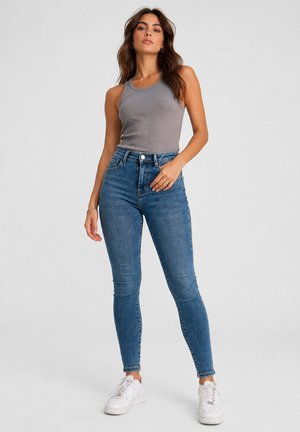 Donna in piedi con maglietta grigia senza maniche, jeans skinny blu a vita alta e sneakers bianche su sfondo uniforme.