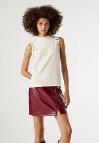 Göndör hajú nő ujjatlan, gombos fehér felsőt visel, és fényes sötétvörös mini szoknyát, amelynek oldalsó zipzárja van, fehér háttér előtt áll.