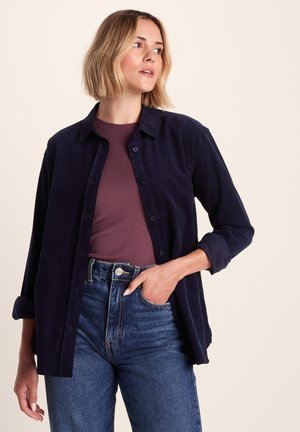 Navy blue corduroy button-up shirt with a collar, long sleeves, and rolled cuffs, worn over a fitted maroon top and high-waisted blue jeans.