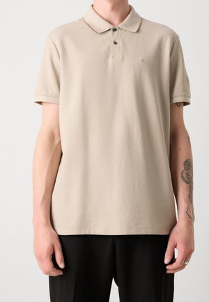 Beige polo shirt in soft fabric with a collar, three buttons, and a subtle brown accent near the collar. Features short sleeves and a relaxed fit.