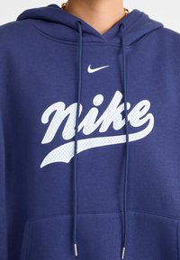 Námořnicky modrá mikina s kapucí z bavlněné směsi s velkým bílým logem "Nike" a malým bílým znakem Swoosh, vybavená šňůrkami a přední kapsou.
