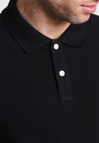 Polo noir en tissu texturé, avec un col et deux boutons blancs au niveau de la patte de boutonnage. Vue rapprochée montrant les détails de couture.