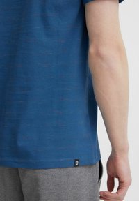 Tricou albastru cu mânecă scurtă, cu un finisaj texturat și fulgi roșii subtili. Dispune de un tiv drept și o etichetă mică cu logo-ul brandului la bază.