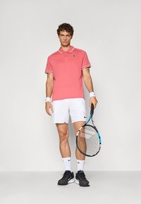 Rosa pikétröja med vit krage och vita ärmdekorationer, kombinerad med vita shorts. Svarta träningsskor, håller i en tennisracket.