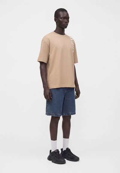 Homme debout portant un t-shirt beige oversize, un short en jean bleu, des chaussettes blanches et des baskets noires sur un fond clair uni.