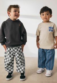 Zwei Jungen stehen nebeneinander. Der eine trägt einen dunkelgrauen Kapuzenpullover und schwarz-weiß karierte Hosen. Der andere hat ein beiges Shirt und blau gestreifte Hosen an.