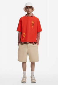 Crvena pamučna majica s vezenim cvijećem, nošena preko ružičaste majice s dugim rukavima. Bež kratke hlače do koljena i bijela kapa s širokim obodom.