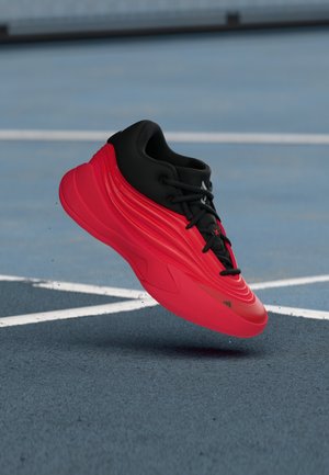 Rød og sort atletisk sneakers i luften over en blå sportsbane med hvide linjer, der fremhæver skodesign og såldetaljer.