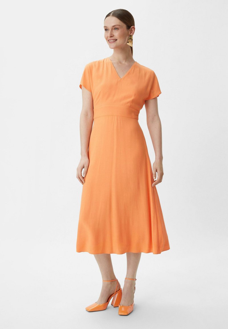 Pomarańczowa sukienka midi z krótkimi rękawami, dekoltem w kształcie litery V i dopasowaną talią. Gładka tekstura oraz sylwetka w kształcie litery A, zestawiona z pasującymi, pomarańczowymi szpilkami.