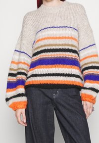 Pull en tricot avec des rayures horizontales dans des tons de beige, orange, noir, violet et vert ; coupe oversized et poignets côtelés.