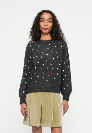 Femme aux cheveux bouclés portant un pull gris foncé avec des motifs de coeurs blancs et rouges, et une jupe plissée en velours côtelé beige.