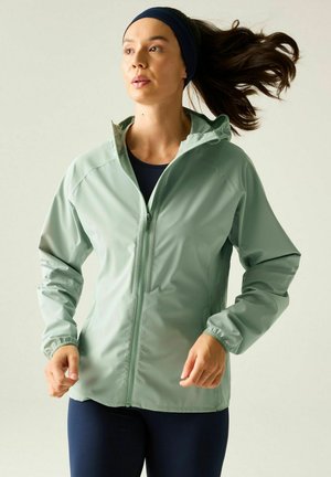 Woman with dark hair in a headband wearing a light green zip-up jacket and navy leggings, in a mid-motion running pose against a neutral background.