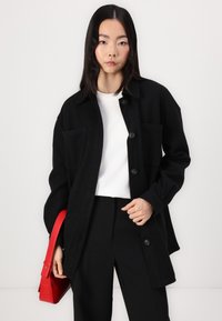 Zwarte oversized jas met een kraag, knoopsluiting aan de voorkant en twee voorzakken. Draagt over een witte shirt en gecombineerd met zwarte broek. Rode handtas als accent.
