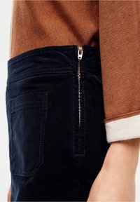 Gros plan d'une personne portant un pantalon en velours côtelé bleu marine avec une fermeture éclair latérale et un haut marron à manches longues avec un revers blanc.