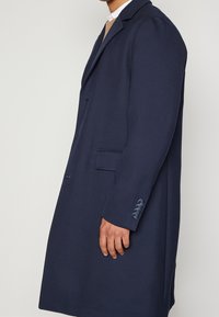 Marinblå kappa med en strukturerad design, lång skärning, texturerad tyg och knapppdetaljer på ärmen; en enda ficka fram.