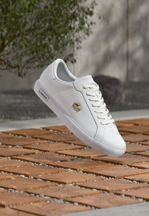 Balta Lacoste kedas ar zeltainu krokodila logotipu, baltas šņorītes un zīmola zoles detaļa, novietots virs terakotas flīžu grīdas ārpus telpām.