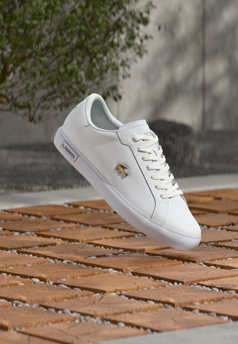 Άσπρο αθλητικό παπούτσι Lacoste με χρυσό λογότυπο κροκόδειλου, άσπρα κορδόνια και λεπτομέρεια σε σόλα με εμπορικό σήμα, τοποθετημένο πάνω σε πλακόστρωτο δάπεδο σε εξωτερικό χώρο.
