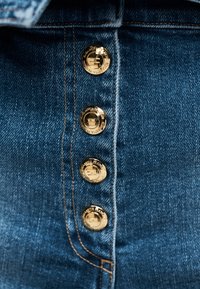 Κοντινή λήψη μπλε υφάσματος τζιν με τέσσερα λαμπερά χρυσά κουμπιά τοποθετημένα κάθετα κατά μήκος ραμμένης ραφής.