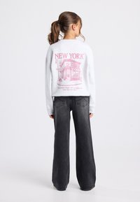 Tyttö, jolla on ruskea poninhäntä, kääntyy poispäin, päällään valkoinen huppari, jossa on vaaleanpunainen New Yorkin ja bagel-kaupan tulostus, sekä löysät mustat farkut.