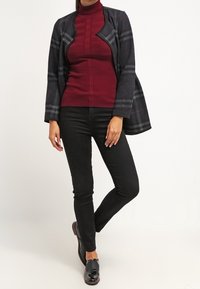 Jeans neri aderenti, dolcevita burgundy a coste e un cappotto avvolgente a quadri scuri con bordi frontali curvi e design aperto, indossato con scarpe nere.