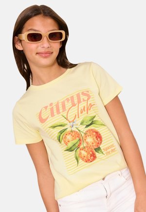 Jonge vrouw die beige rechthoekige zonnebrillen draagt en een geel t-shirt met "Citrus Club" en een oranje fruitgrafiek, gecombineerd met een witte broek.