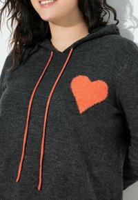 Sweat à capuche gris avec cordons de serrage orange et un cœur en peluche orange sur le côté gauche de la poitrine, présentant une texture douce et un design décontracté.