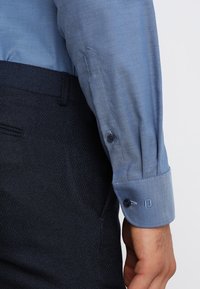 Lichtblauw, gestructureerd overhemd met knoopmanchetten, gecombineerd met een donkere broek. Heeft een gladde afwerking en contrasterende knoopdetails.