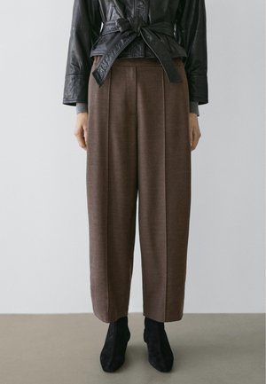 Woman wearing brown high-waist wide-leg trousers with front pleats, black ankle boots, and a belted black leather jacket.