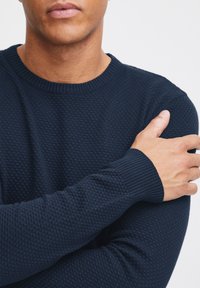 Navy blue knit sweater with a textured honeycomb pattern, ribbed crew neckline, and cuffs, worn with arms crossed.