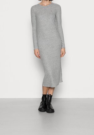 Personne portant une robe longue moulante en maille côtelée grise à manches longues et des bottines noires, debout devant un fond uni.