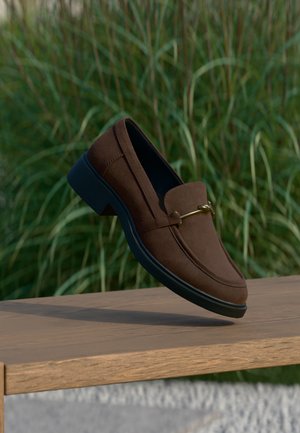 Mocassin en daim marron avec semelle noire et détail métallique doré, en équilibre sur un banc en bois avec un fond d’herbe verte.