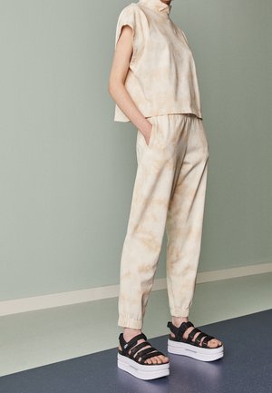 Personne portant un pantalon tie-dye beige clair et un haut sans manches avec des sandales à plateforme noires, debout contre un mur vert pâle.