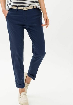 Kobieta ubrana w granatowe spodnie z mankietami, beżowy pasek, pasiasty top i beżowe wsuwane buty, stojąca z rękami w kieszeniach.