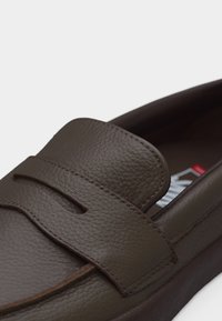 Σκούρο καφέ δερμάτινο μοκασίνι με στρογγυλεμένη μύτη και ραμμένο λουράκι τύπου penny πάνω από το πάνω μέρος του παπουτσιού, εμφανίζεται σε κοντινή λήψη.