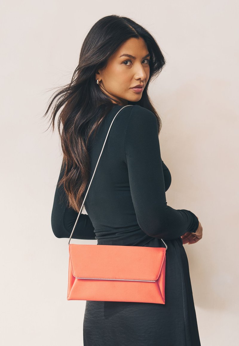 Sac pochette corail à texture lisse, design à rabat, avec une chaîne argentée en bandoulière, porté sur un haut noir à manches longues.