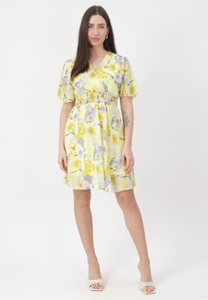 Mujer de pie con vestido corto amarillo con estampado floral, cintura fruncida y sandalias blancas de tacón sobre un fondo blanco.