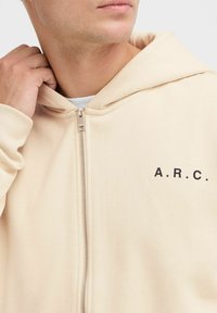 Beige Zip-Up-Hoodie mit lockerer Passform, ausgestattet mit einer Kordelzugkapuze und schwarzem "A.R.C."-Text auf der Brust. Glatte Stofftextur.