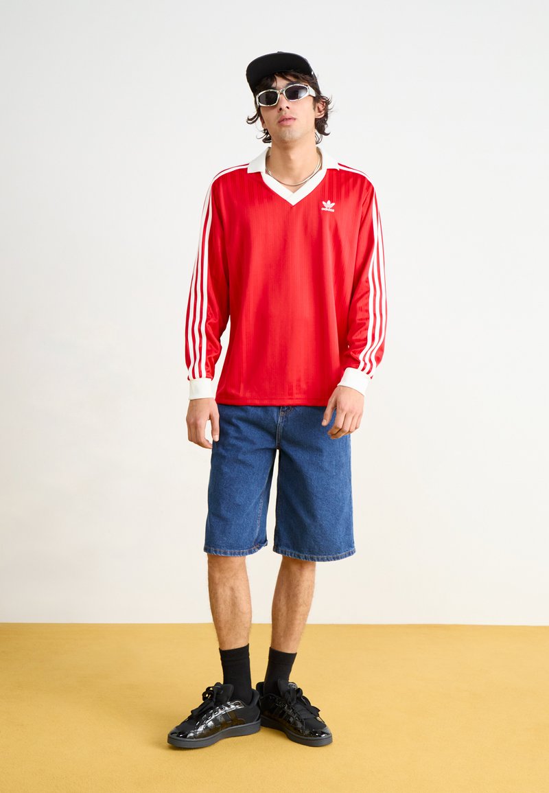 Jersey rojo de manga larga con cuello blanco y tres rayas blancas en las mangas, combinado con pantalones cortos de denim azules y zapatillas negras.