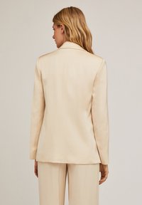 Blazer bej, croșetat, cu o textură netedă, rever tăiat și două fante la spate. Puratat împreună cu pantaloni asortați, într-o ținută coordonată.