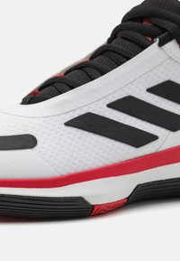 Scarpa sportiva bianca con tomaia testurizzata, strisce nere e accenti rossi sulla suola. I lacci sono neri, con un design elegante.