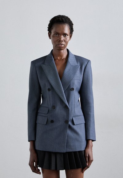 Blazer croisé en pinstripe bleu marine, doté de grands revers, de quatre poches frontales et de boutons noirs contrastés, associé à une jupe plissée noire.