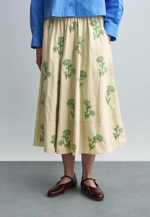 Modrá košeľa s dlhým rukávom zapnutá v béžovej midi sukni so zelenou kvetinovou výšivkou, kombinovaná s tmavohnedými topánkami Mary Jane na sivom podlahe.