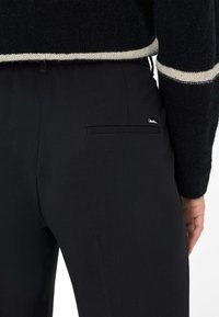 Pantalons noirs ajustés avec une texture lisse, comportant une poche arrière et un accent subtil avec un logo. Associés à un pull en tricot sombre.